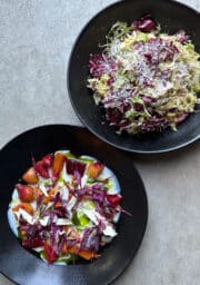 Two black plates of food on a gray surface. The top plate contains a salad with grated cheese. The bottom plate has a dish with colorful vegetables and white sauce, garnished with red leaves.