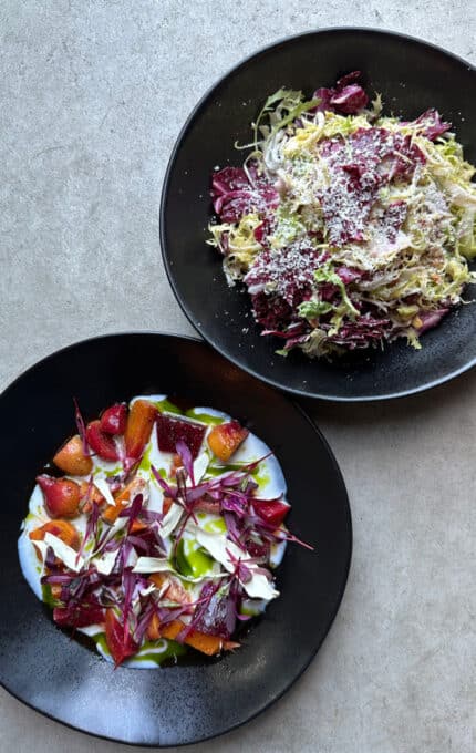 Two black plates of food on a gray surface. The top plate contains a salad with grated cheese. The bottom plate has a dish with colorful vegetables and white sauce, garnished with red leaves.