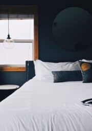 A modern bedroom with a neatly made bed featuring white linens and decorative pillows. A round mirror hangs on a dark wall. A small side table with a hanging light bulb is near the window, which has partially drawn curtains.