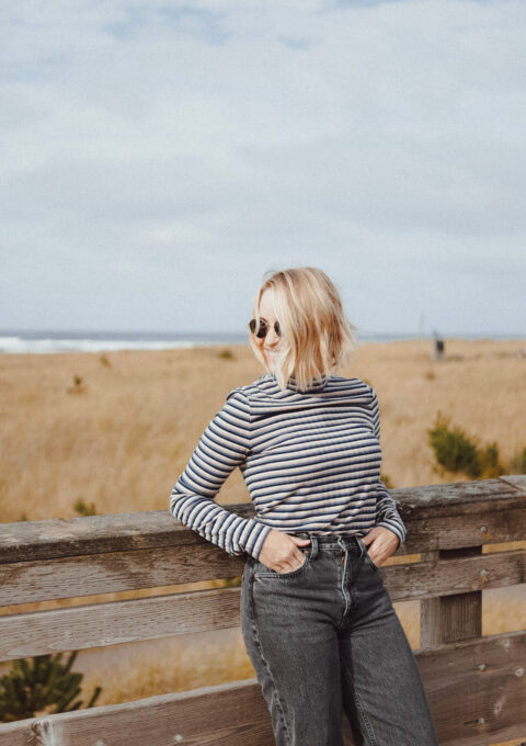 A person with blonde hair wearing sunglasses, a striped long-sleeve shirt, and high-waisted black jeans leans against a wooden fence. There is a grassy field and the ocean in the background under a cloudy sky.