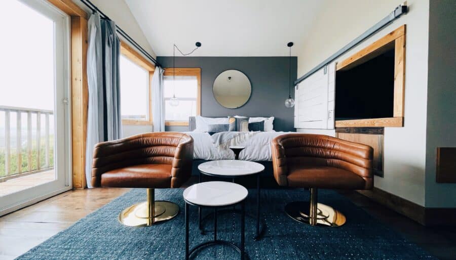 A modern bedroom features a bed with gray bedding against a dark accent wall. Two brown leather chairs and a set of three round tables are on a dark rug. Large windows with curtains flank the sides, and a circular mirror hangs above the bed.