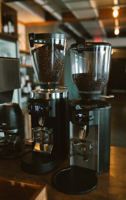 Two modern coffee grinders with digital displays are filled with coffee beans and placed side by side on a wooden counter in a coffee shop setting.