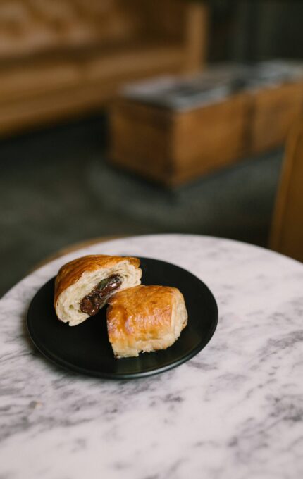 A black plate with two chocolate croissants is placed on a round marble table. The background features a brown couch and a wooden chest, slightly out of focus.