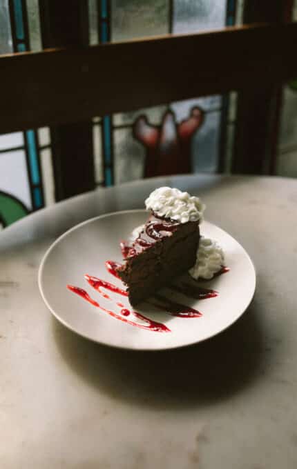 A slice of chocolate cake on a white plate is topped with whipped cream and drizzled with red sauce. The plate is set on a marble surface, with a stained glass window in the background.