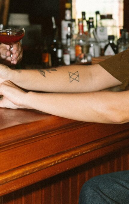 Two people sit at a bar, each holding a cocktail glass. One person has long hair and visible tattoos on their arm. Various bottles are visible in the background behind the bar.