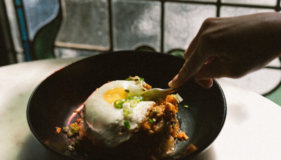 A hand is using a spoon to scoop food from a black bowl. The dish contains rice topped with a fried egg and garnished with green onions, placed on a white surface with a stained glass window in the background.