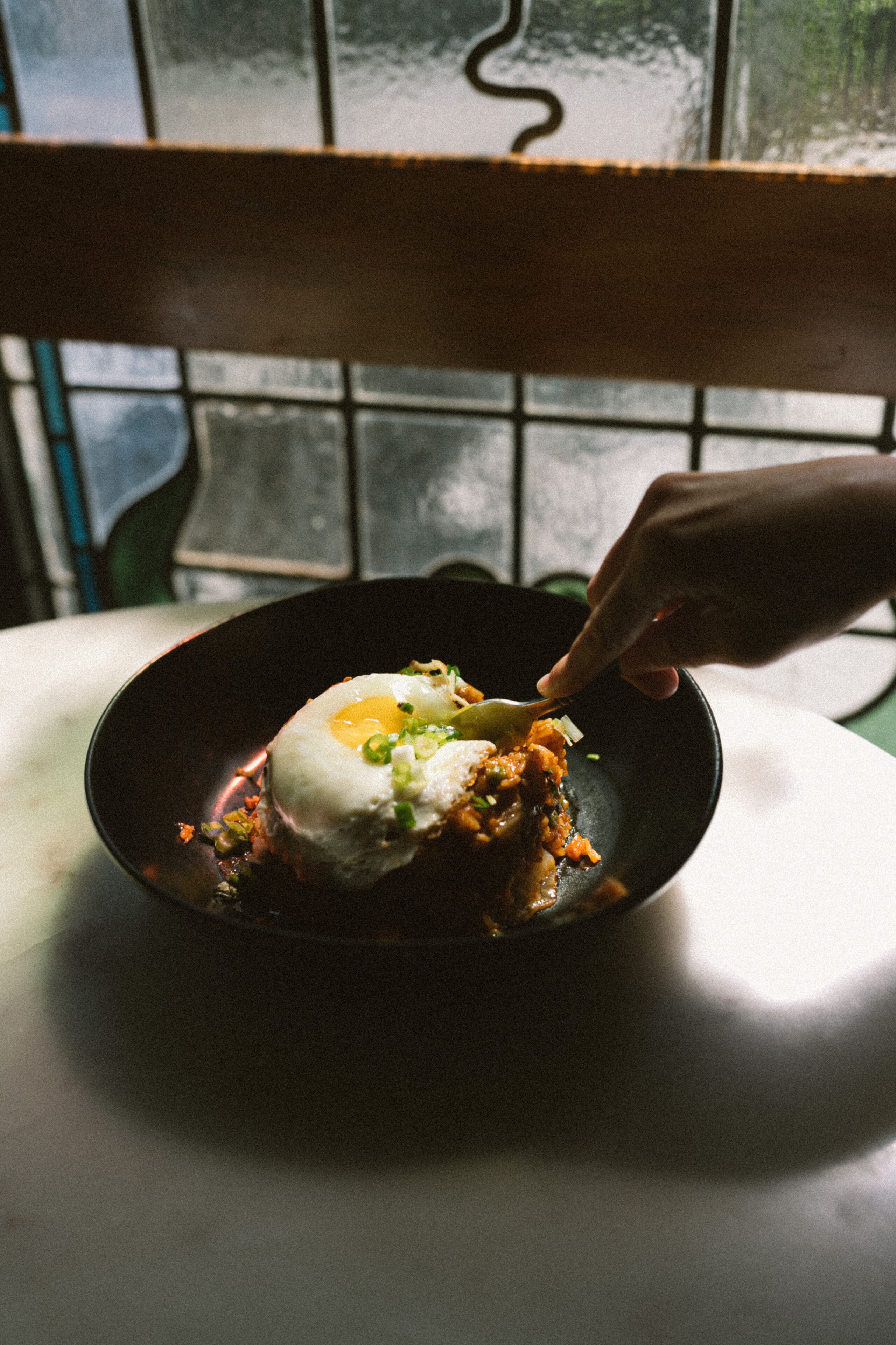 A hand is using a spoon to scoop food from a black bowl. The dish contains rice topped with a fried egg and garnished with green onions, placed on a white surface with a stained glass window in the background.