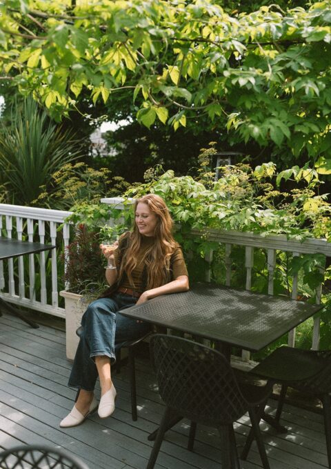 A woman sits alone at an outdoor table on a patio surrounded by green plants. She holds a glass and smiles, with empty chairs and tables nearby under leafy branches.