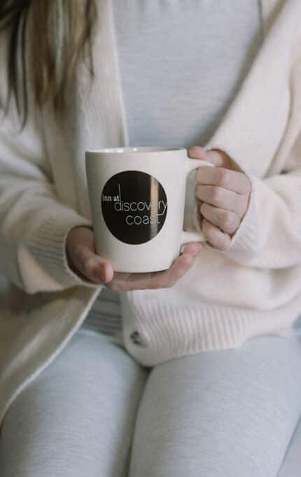 A person wearing light-colored clothes sits on a bed, holding a white mug with a black circle and text that reads "discovery coast." Their face is not visible.