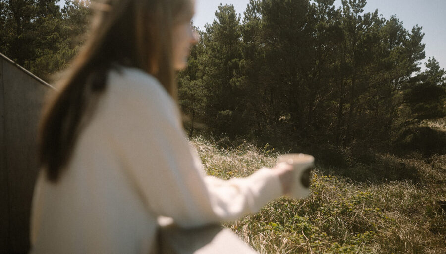 A person with long hair and a light sweater holds a mug while standing outdoors near a railing, overlooking a grassy area with trees in the background. The photo is slightly out of focus.