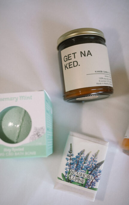 A jar labeled "GET NAKED," a box of rosemary mint bath bomb, and a bar of soap labeled "Seashore Lupine" are arranged on a white surface.