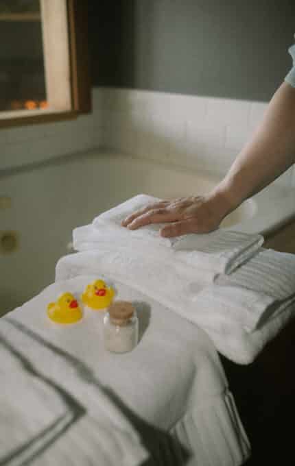 A person places a folded white towel on a stack of towels near a bathtub. Two yellow rubber ducks and a small jar filled with bath salts are on top of the towels.