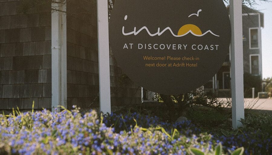 A round sign reads "Inn at Discovery Coast" with instructions to check in at the adjacent Adrift Hotel, surrounded by blue flowers and greenery near a shingled building on a sunny day.