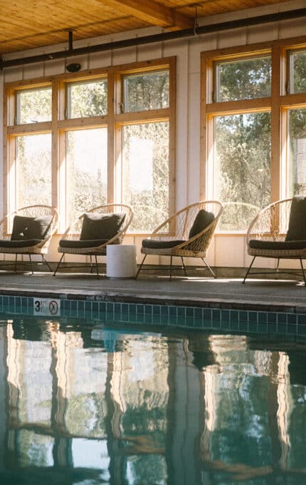 Indoor swimming pool with clear water, surrounded by several wicker lounge chairs with cushions. Large windows let in natural light, and potted plants are placed near the seating area. Wooden ceiling is visible above.