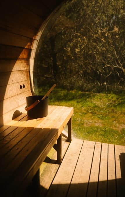 A wooden sauna bench with a black bucket and ladle is shown in sunlight, next to a large window overlooking green grass and trees outside.