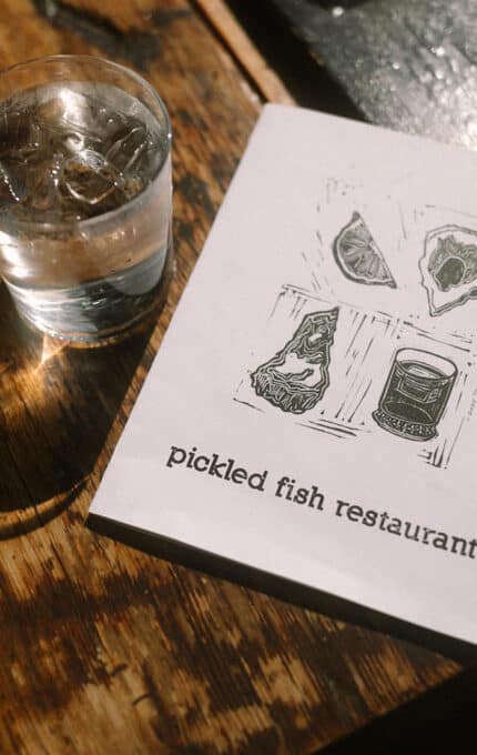 A glass of ice water sits on a wooden table next to a menu for "pickled fish restaurant," which features simple line drawings of oysters and a jar on the cover. Sunlight shines in from a nearby window.