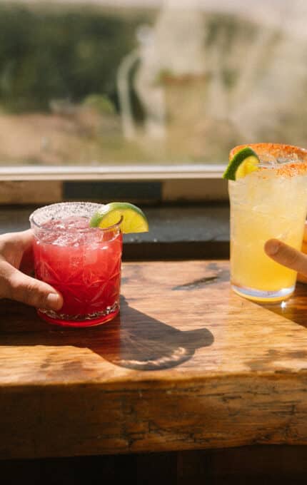 Two people sit at a wooden table by a window, each holding a cocktail; one is red with a lime wedge, and the other is yellow with a chili rim and lime. There are glasses of water in the background.