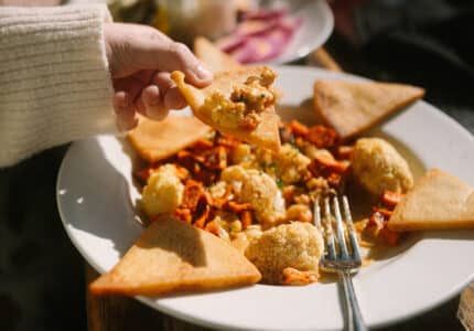 A hand holding a piece of pita bread dips into a plate with roasted cauliflower, diced vegetables, sauce, and more pita triangles. A fork rests on the plate, with another meal and a pink drink blurred in the background.