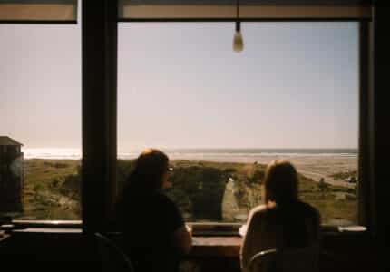 Two people sit at a counter facing a large window with a view of a beach, grassy dunes, and the ocean under a clear sky. Sunlight streams in, and a single hanging light bulb is visible at the top of the window.