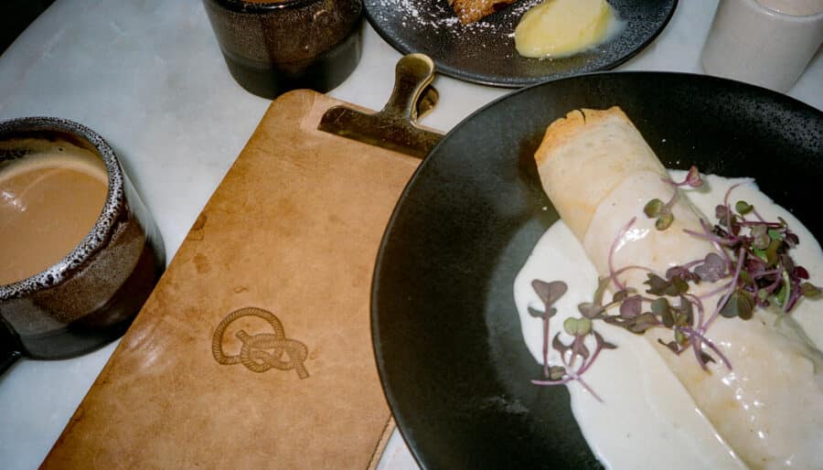 A meal is set on a table, featuring a crepe with cream sauce and microgreens on a black plate, a cup of coffee, a small dessert with powdered sugar and butter, and a brown leather menu with an embossed logo.