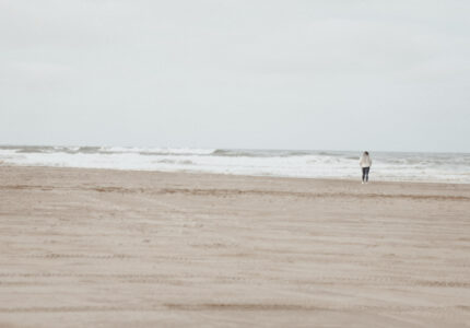 A person wearing a light jacket and jeans walks alone on a vast, empty beach towards the ocean under a cloudy sky. The sand appears untouched, and gentle waves are seen in the distance.