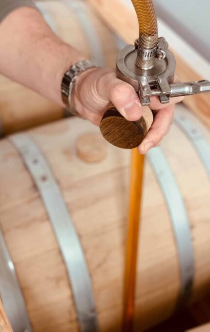 A person is using a hose to fill wooden barrels with a liquid. The person is holding the hose securely, directing the flow into one of the barrels. The barrels have metal bands around them, and the wood appears light in color.