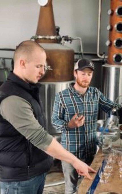 Two men are seen working together in a distillery. One, wearing a black vest, is pointing at a row of glasses on the table, while the other, in a checkered shirt and a cap, observes. Behind them are large distillation tanks and other brewing equipment.