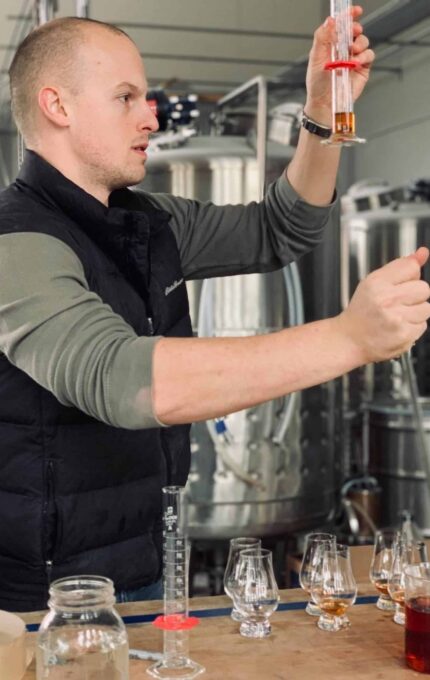 A person in a black vest carefully examines a glass cylinder with a reddish liquid. Several other glass containers with various liquids are on the table. Industrial tanks are visible in the background, suggesting a laboratory or brewing environment.