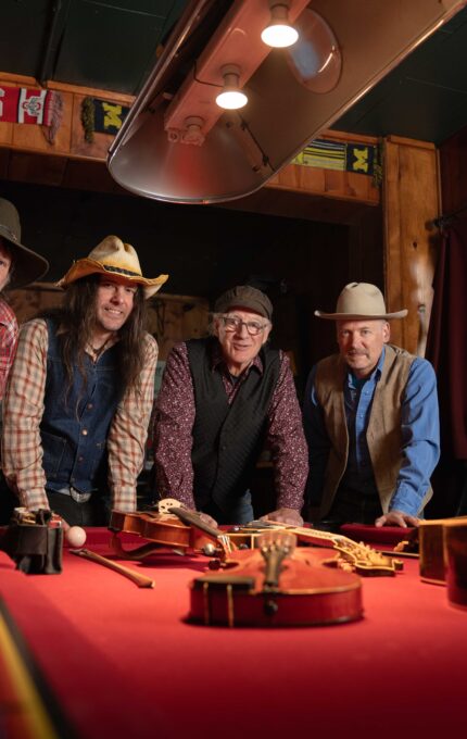 Four men wearing hats and casual attire stand around a pool table with musical instruments, including a guitar and mandolin, atop it. The room has wood-paneled walls with a "Buckeyes" banner. The lighting is warm and focused.