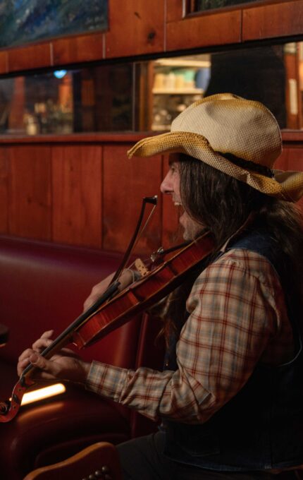 A person wearing a cowboy hat plays a violin while seated in a rustic, wood-paneled restaurant. The dimly lit interior features red leather booths, a polished wooden table, and decorative paintings on the walls.