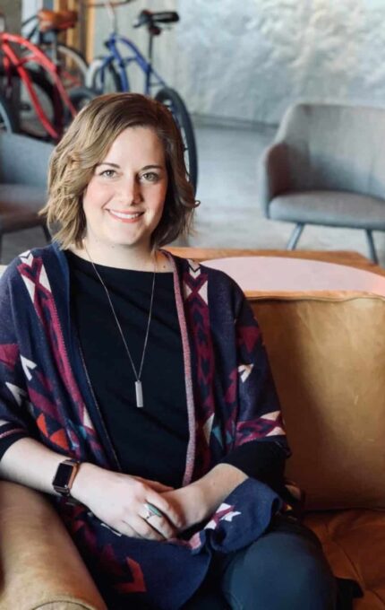 A person with short, wavy hair sits on a tan leather couch in an industrial-style room. They are wearing a patterned shawl, black top, and wristwatch, and are smiling. In the background are two gray chairs and a few bicycles.