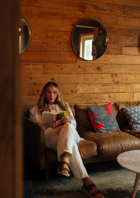 A woman with long blonde hair sits on a brown leather sofa reading a book in a room with wooden walls, round mirrors, and decorative pillows, including one with a red crab design.