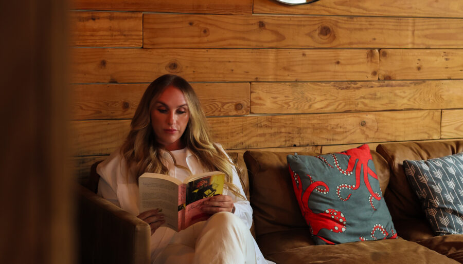 A woman with long blonde hair sits on a brown leather sofa reading a book in a room with wooden walls, round mirrors, and decorative pillows, including one with a red crab design.