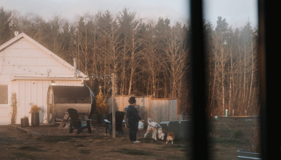 A person stands outside a white house near a metal structure, with trees in the background and two dogs nearby. The image is partially obscured by a dark object in the foreground.