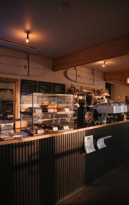 A coffee shop counter with a display case of pastries, a large espresso machine, and menus on the wall; surfboards are mounted as decor above the counter. Warm lighting fills the space.
