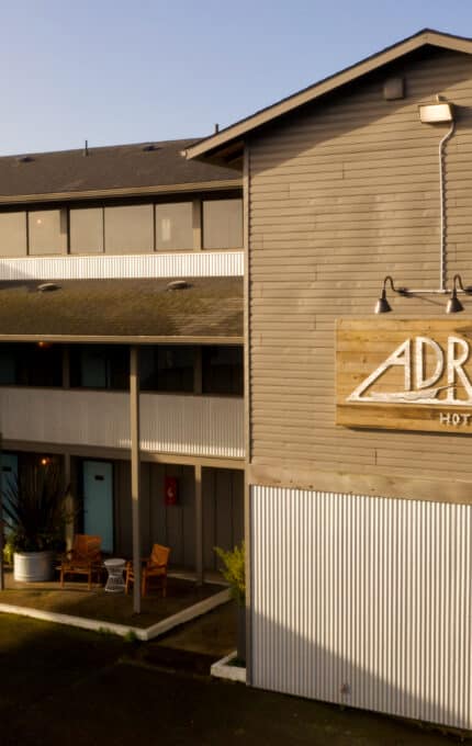 A three-story building with a wooden exterior and a sign reading "Adrift Hotel" is shown. The structure has balconies, outdoor seating, and a flat driveway. The sky is clear, and there is a glimpse of the sea in the background.