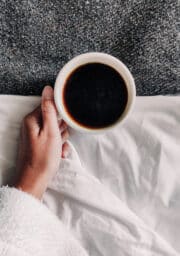 A person holds a cup of black coffee in a white mug, partially covered by a white blanket. The background features a textured, dark gray surface resembling carpet. The person is wearing a white robe.