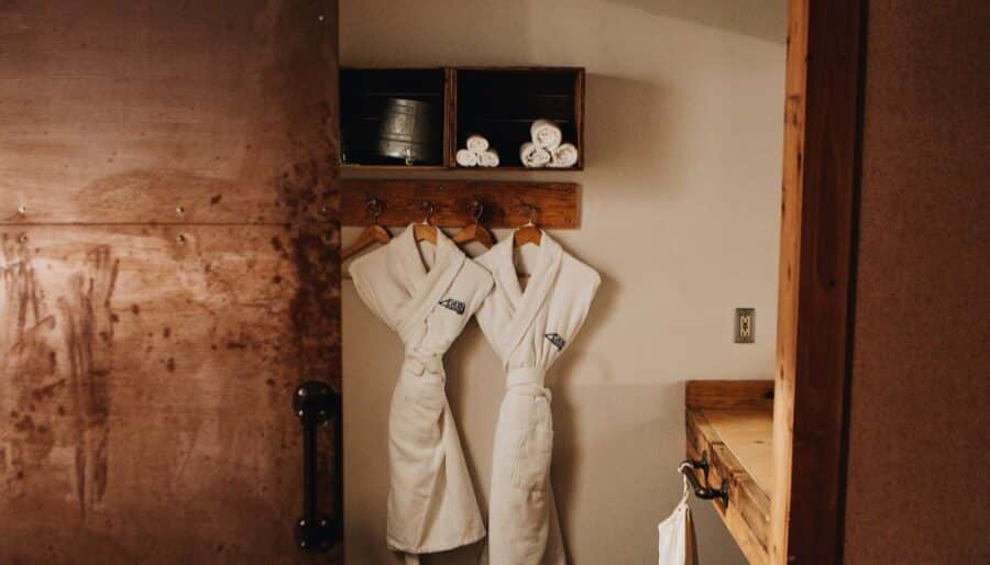 Two white bathrobes hang side by side on wooden hooks in a rustic room. Above them, shelves hold rolled white towels and a metal bucket. A partially open wooden door is visible on the left side of the image.