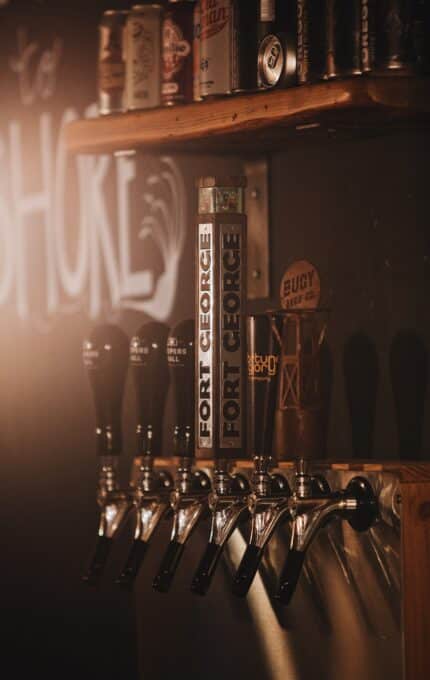 A dimly lit bar features six beer taps mounted on a wooden panel. The central tap is labeled "Fort George." Shelves above the taps display assorted beer cans and glasses, with a chalkboard wall in the background.