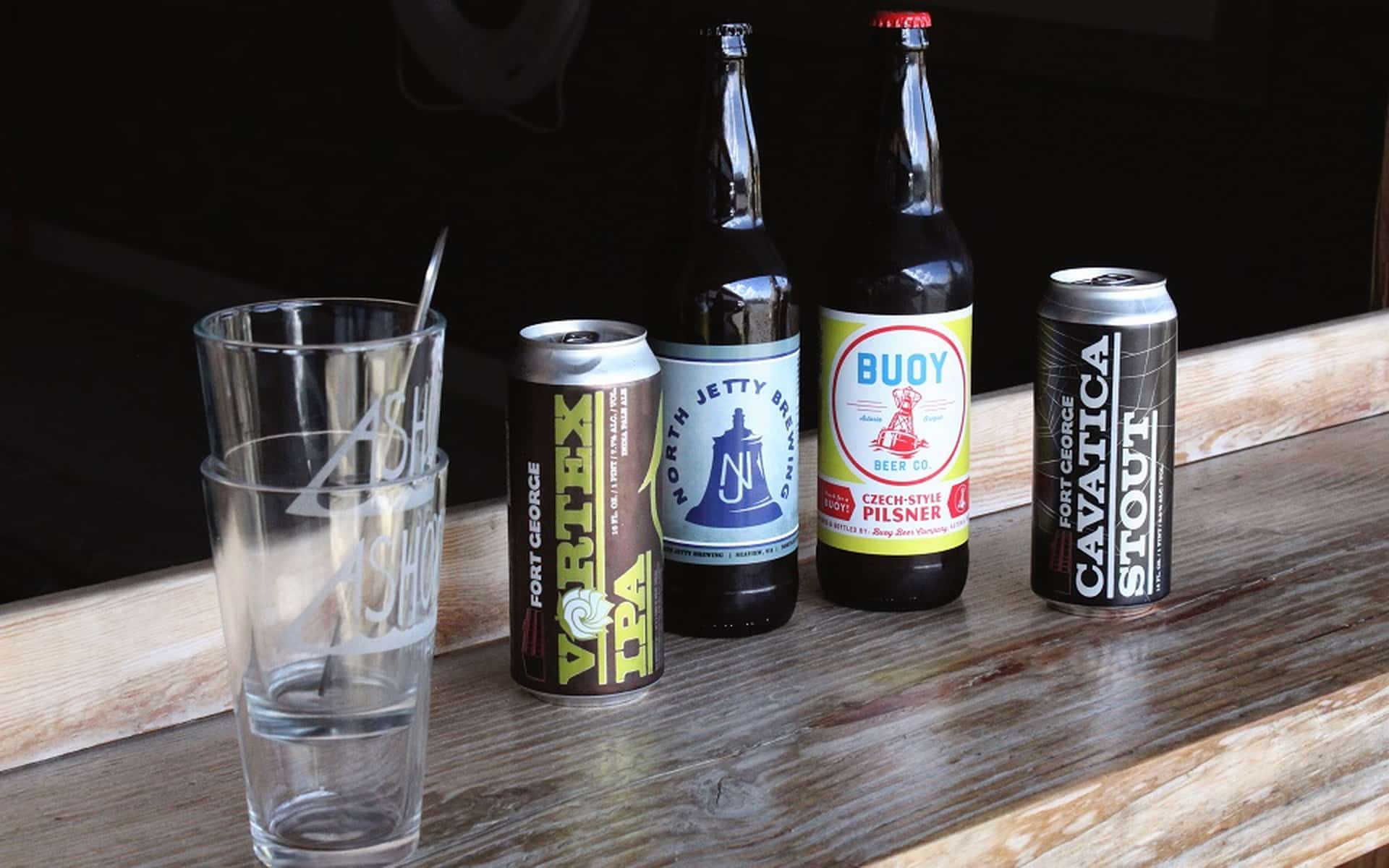 Four different beer bottles and cans are displayed on a wooden surface next to two empty glasses bearing the logo of "Astoria Brewing Company." From left to right, drinks include a Vortex IPA, North Jetty Brew, Buoy Czech-Style Pilsner, and Cavicatia Stout.