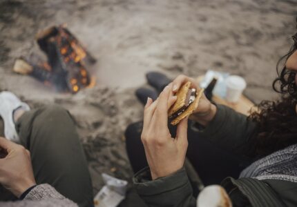 Two people sit on the sand near a small campfire on a beach. One person holds a s'more in their hands, with melted chocolate and marshmallow visible. The scene includes beach sand, logs in the fire, and some scattered items around them.