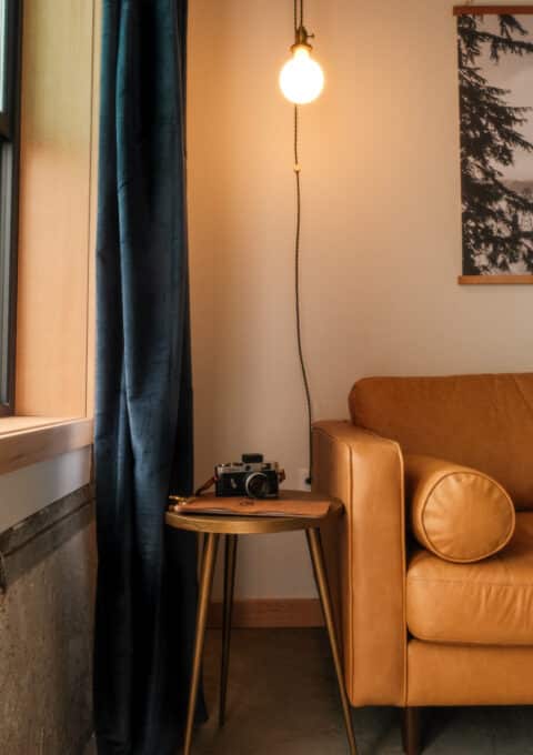 A cozy corner with a tan leather sofa next to a small wooden side table. A vintage camera sits on the table. A single bulb hangs from the ceiling, casting warm light. A dark curtain and a framed picture are on the wall nearby.