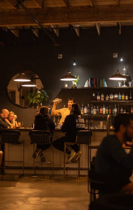 A group of people sit and talk at a bar in a warmly lit, modern interior with exposed beams. Shelves with bottles and glasses are behind the bar, and other patrons are seated at tables in the foreground.