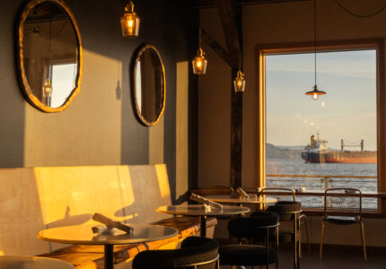 Sunlit café interior with empty tables and chairs, round mirrors on a dark wall, and pendant lights. A large window shows a cargo ship on the water and distant mountains outside.