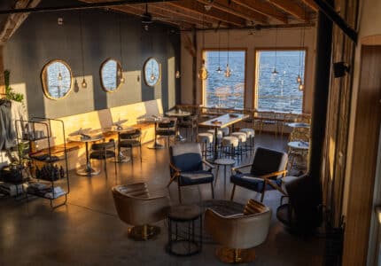 A modern restaurant interior with wooden floors, round and rectangular tables, black and tan chairs, pendant lights, round mirrors on a dark wall, and large windows overlooking a body of water at sunset.