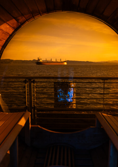 View from inside a wooden barrel sauna looking out to the ocean at sunset, with a cargo ship visible on the water and an orange-tinted sky reflecting through the circular glass window.