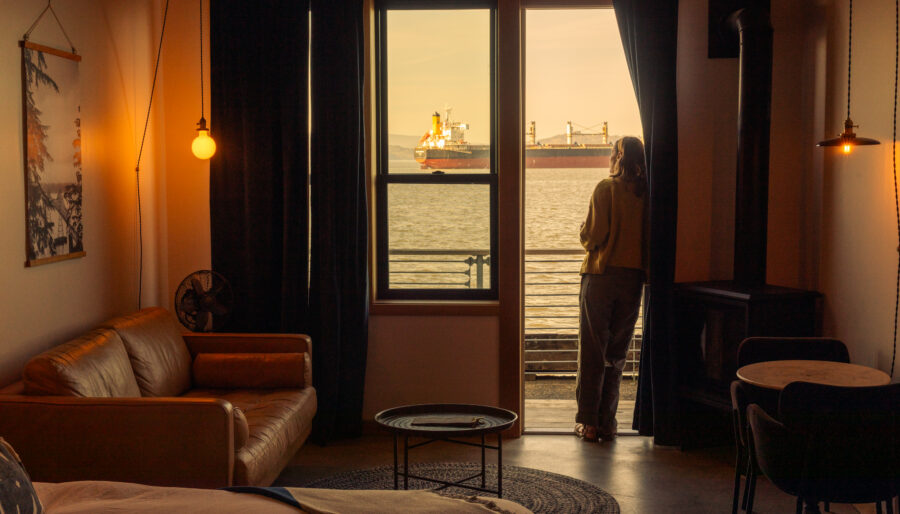 A person stands on a balcony looking out at a large cargo ship on the water, seen from inside a warmly lit living space with a sofa, table, bed, and dark curtains framing the window and door.