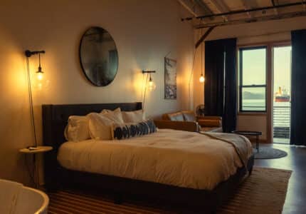 A warmly lit bedroom features a large bed with white bedding, a round mirror above the headboard, hanging light bulbs, a brown leather sofa, and a window with a view of water and a distant ship outside.