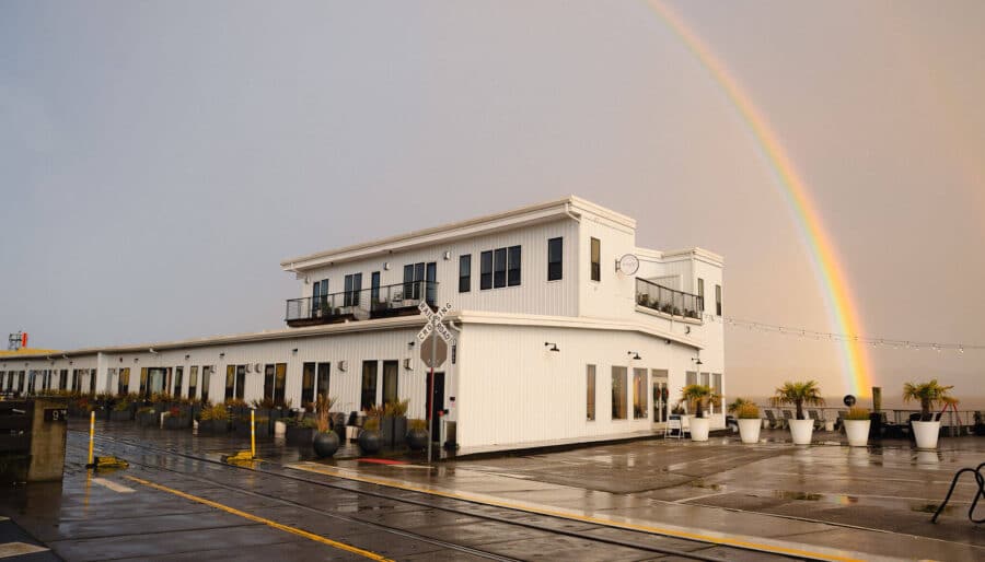 A white two-story building sits on a wet pier with potted plants outside and a rainbow in the sky behind it. The pavement and building reflect light from a recent rain.