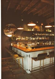 A cozy bar interior with a wooden countertop and several stools. Bottles of various drinks line the shelves on the dark wall. Three round pendant lights hang above the bar, and a circular mirror reflects part of the room.